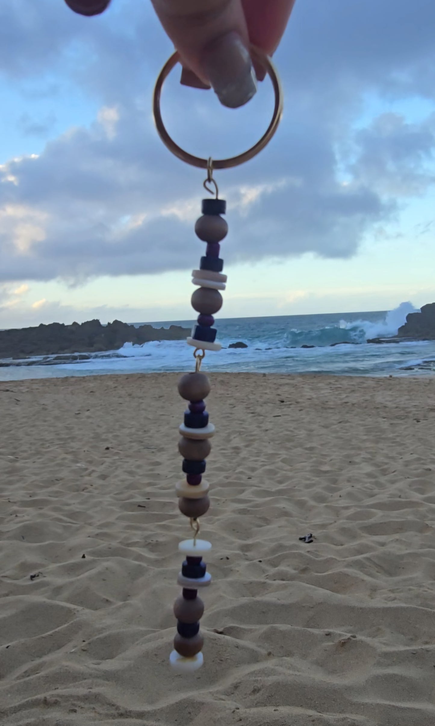 sandy beach with waves crashing on the rocks. Well-manicured hands holding a customed made beaded bracelet