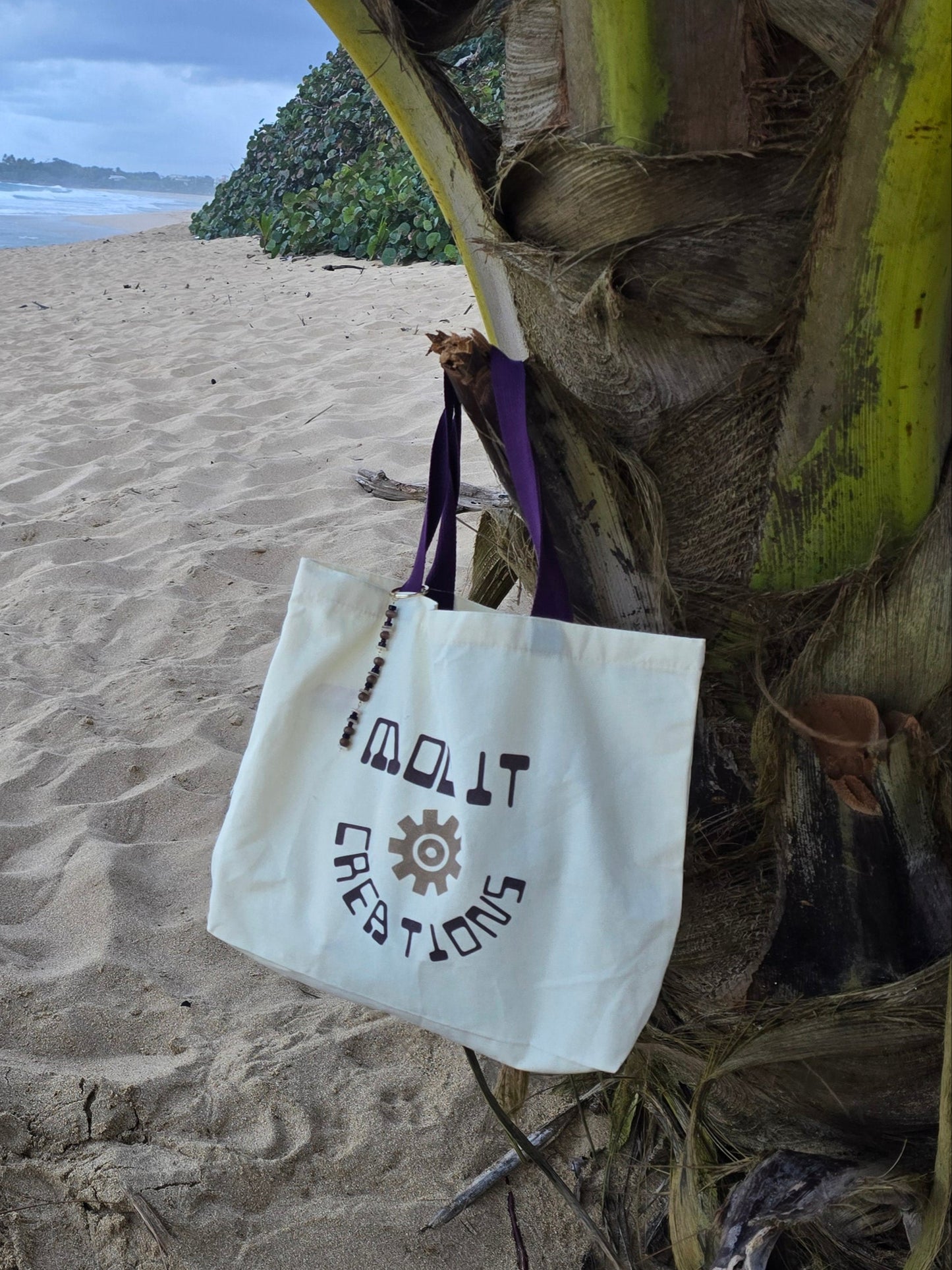 Sandy beach with Mood it creations logo tote bag hanging on a palm tree.